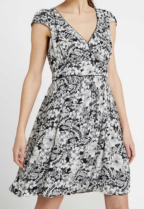 Žena v černobílých květinových šatech s krátkým rukávem a výstřihem do V, přiléhavých v pase a rozšířených v sukni.