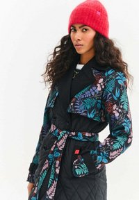 Prešívaný čierny trenčkot s výraznými kvetinovými vzormi v modrej, ružovej a zelenej farbe. Má široký golier, dvojradové zapínanie a opasok v páse.