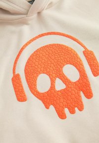 Sudadera crema con un gráfico de calavera naranja texturizado con auriculares. El diseño presenta un patrón agrietado y un fuerte contraste.