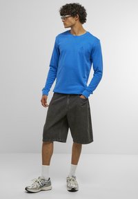 Felpa blu a maniche lunghe con collo a giro, abbinata a pantaloni denim neri. Il modello indossa scarpe da ginnastica e calze bianche a girocchi.