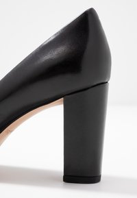 Chaussure à talon haut noire avec une finition brillante, présentant un talon carré épais et un bout pointu, affichant une texture de cuir lisse.