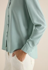 Personne portant une chemise boutonnée bleu clair avec les manches longues, la main posée de manière décontractée dans la poche avant d'un pantalon blanc cassé, devant un fond neutre.