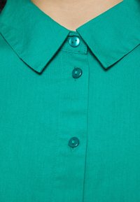 Žalia, apykaklė su minkšta tekstūra, turi tris derančius mygtukus priekyje. Audinys atrodo lengvas ir lygus.