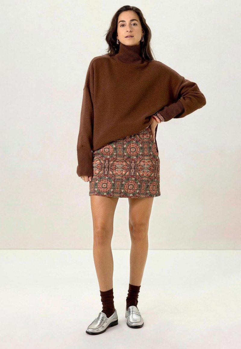 Pull surdimensionné marron à col roulé, jupe mini à motifs circulaires, mocassins argentés et chaussettes marron foncé. Arrière-plan clair.