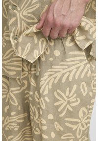 Beežist kangast särk suurte helekollaste lillemustritega. Krae juures näha nuppe; esitasku on osaliselt avatud ja sellel on paeladetail.