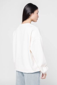 Sudadera blanca de gran tamaño con hombros caídos, textura de tela suave y cuello redondo. Pantalones de pana azules son parcialmente visibles.