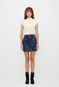 Gonna mini in ecopelle blu navy con fronte diagonale, dettagli dorati e zip laterale. Top corto a maniche corte in avorio con collo alto. Stivali alla caviglia neri.