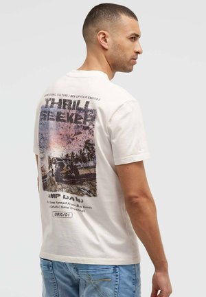 Man wearing white t-shirt with off-road vehicle and "Thrill Seeker" text on the back, paired with light blue jeans.