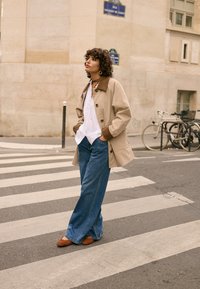 Kobieta o kręconych włosach, ubrana w beżowy płaszcz i dżinsy, stoi na miejskim przejściu dla pieszych obok zaparkowanych rowerów.