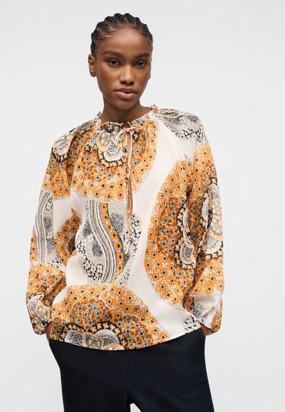Femme portant un chemisier ample à manches longues avec des motifs floraux et cachemire orange, blanc et noir, associé à un pantalon foncé, debout les mains dans les poches.