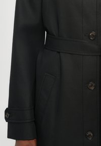 Melns mētelis ar pieguļošu dizainu, jostu viduklī un noapaļotu apkakli. Ievērojami pogas, sānu kabatas un uzvelkami piedurkne ar pogu detaļām.