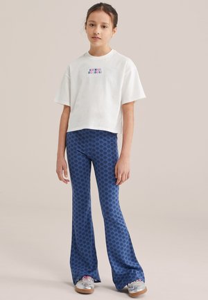 Tânăr purtând un tricou alb scurt cu text și pantaloni evazați albaștri cu model, stând pe un fundal bej simplu.