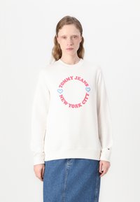 Kremgenser med ribbet krage og mansjetter. Har rød og blå tekst i et sirkulært mønster: "TOMMY JEANS NEW YORK CITY" med hjerteaksenter.