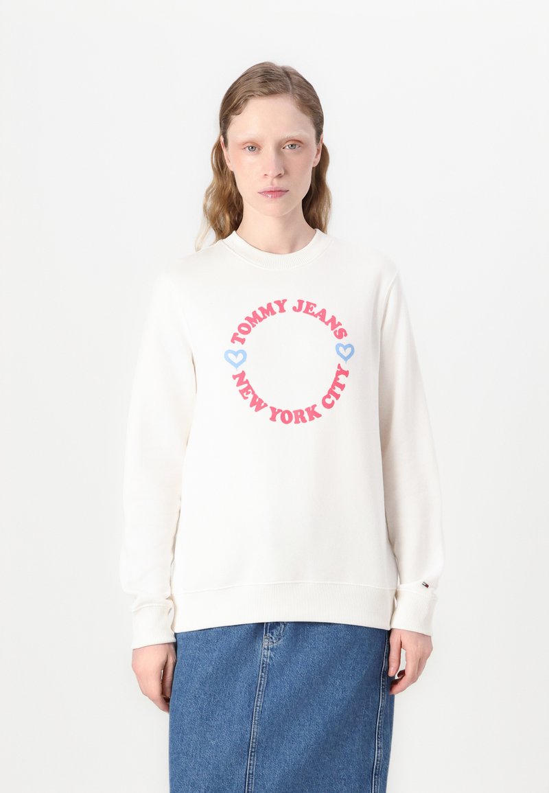Kremgenser med ribbet krage og mansjetter. Har rød og blå tekst i et sirkulært mønster: "TOMMY JEANS NEW YORK CITY" med hjerteaksenter.