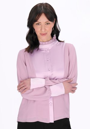 Žena s tmavými vlnitými vlasy, oblečená v světle fialové blůze s volánovým límcem a manžetami, s překříženýma rukama, na bílém pozadí.