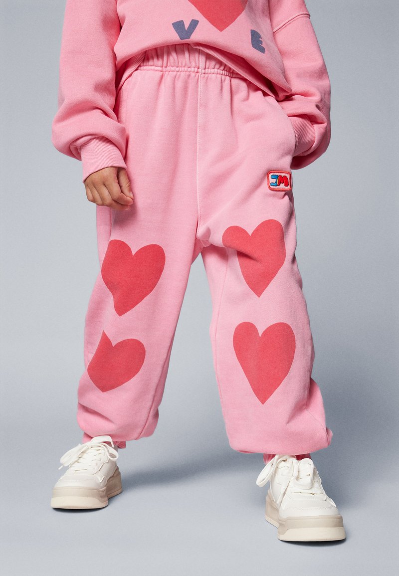 Rózsaszín melegítőnadrág nagy piros szívmintákkal, puha pamut anyagból, rugalmas derekú, oldalán logóval, fehér sportcipővel párosítva.