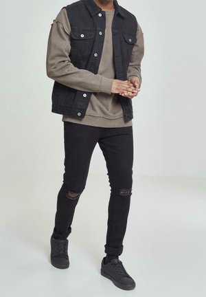 Czarna kamizelka dżinsowa na szarym, oversize'owym swetrze, w połączeniu z podartymi czarnymi jeansami i czarnymi trampkami. Jednolite tło.