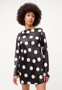 Rochie din satin negru cu buline albe mari, mâneci lungi și gât rotund, cu o textură netedă și fante laterale.