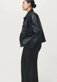 Donna con capelli raccolti in un bun, che indossa una giacca in pelle nera oversize e pantaloni a gamba larga, posata di profilo su uno sfondo semplice.