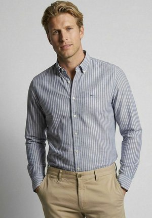 Blond man wearing a gray and white striped button-up shirt and beige pants, standing with hands in pockets against a plain background.