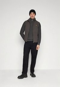 Gilet grigio scuro sopra un maglione nero, pantaloni neri e scarpe nere; dettaglio a maglia testurizzata sul maglione; completato da un berretto nero.