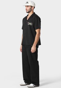 Svart kortermet skjorte med knapper og brodert "PAS DE MONACO" tekst, sammen med svarte vide bukser og hvite joggesko. Nøytral caps brukt.