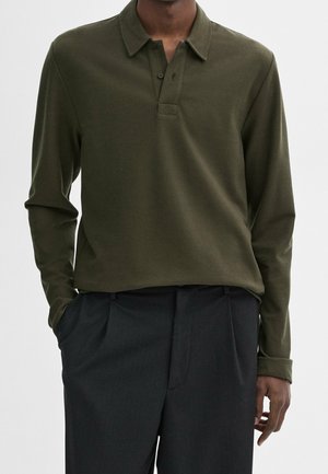 Mężczyzna w oliwkowo-zielonej koszulce polo z długim rękawem, włożonej do czarnych spodni, z rękami w kieszeniach na jednolitym tle.