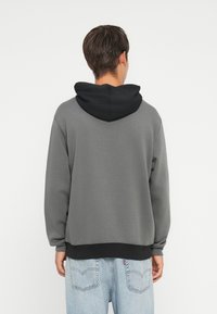 Hanorac gri cu glugă neagră, guler rotund și tiv striat. Materialul pare moale și texturat. Design casual cu umeri lăsați.