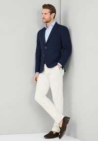 Blazer blu navy, camicia a righe chiare, pantaloni bianchi, mocassini marroni. Il blazer è dotato di due bottoni e di una tasca frontale. Taglio liscio e su misura nel complesso.