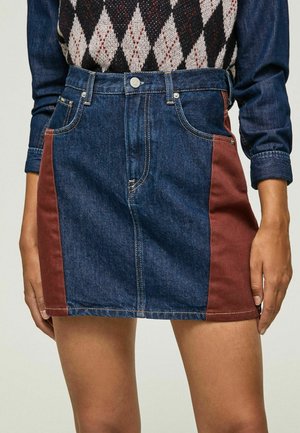 Femme portant une mini-jupe en jean bleu avec des panneaux latéraux couleur rouille et une chemise à manches longues bleu foncé avec un gilet à motif losange.