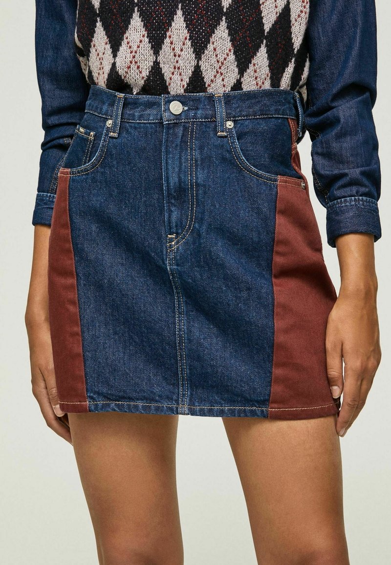 Femme portant une mini-jupe en jean bleu avec des panneaux latéraux couleur rouille et une chemise à manches longues bleu foncé avec un gilet à motif losange.