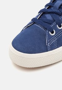 Blå mockaskor med en texturerad vit gummisula, med dubbla sömnadsdetaljer och runda metallöglor för snörning.