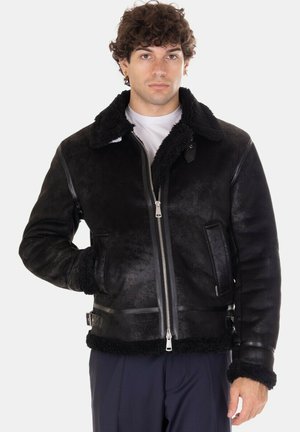 Uomo con giacca nera in shearling con colletto di pelliccia, chiusura frontale con cerniera, fibbie laterali e pantaloni scuri su sfondo semplice.