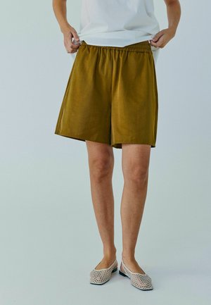 Persoon die olijfgroene korte broek met elastische taille aanpast, gekleed in een wit bovenstuk en witte instapmesh schoenen, staand op een lichtgrijze vloer.