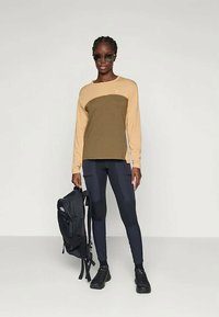 Άτομο που φορά γυαλιά ηλίου, μπεζ και ελιά πράσινο μακρυμάνικο πουκάμισο, μαύρες κολάν και μαύρα παπούτσια, κρατώντας μια μαύρη τσάντα πλάτης στο πλάι.