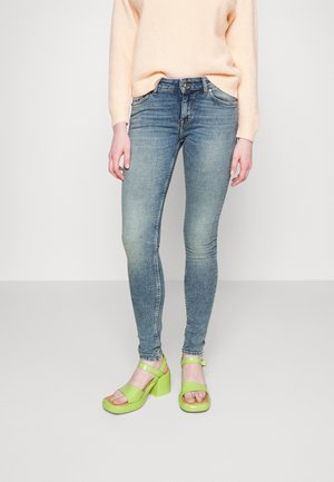 Vaqueros ajustados de denim azul combinados con sandalias de tacón bloque en color verde lima. Los vaqueros presentan un ligero desgaste y una cintura de altura media.