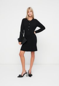 Μαύρο εφαρμοστό φόρεμα με μακριά μανίκια, κλείσιμο με κουμπιά και φαρδιά βάση. Συνδυάζεται με μαύρες γόβες slingback με διακόσμηση φιόγκου.