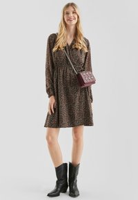Blumendruckkleid in Dunkelbraun mit langen Ärmeln, gerafftem Taillenbereich und knieumspielendem Saum. Kombiniert mit einer kleinen burgunderfarbenen Handtasche und schwarzen Stiefeln.