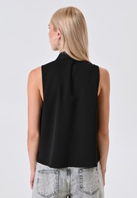 Una donna con lunghi capelli biondi indossa un top nero senza maniche e jeans grigio chiaro, vista di spalle su uno sfondo semplice.