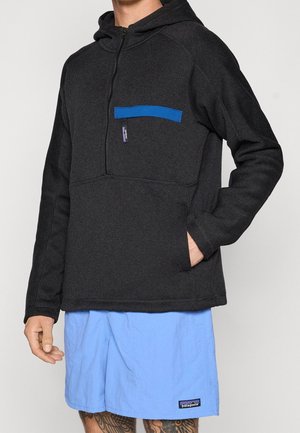 Persoon die een zwarte capuchontrui draagt met een blauw borstzakje en lichtblauwe korte broek met een merklabel op de zoom.