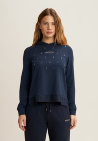 Felpa blu navy con vestibilità ampia, caratterizzata da un logo frontale e borchie decorative. Tessuto morbido, maniche lunghe e dettaglio con cordoncino all'orlo.