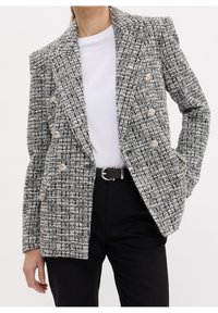 Blazer croisé en tweed noir et blanc avec des boutons argentés, porté sur une chemise blanche et un pantalon noir avec une ceinture noire.