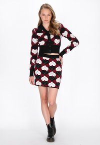 Pletený dvojdielny outfit pozostávajúci z čierneho skráteného svetra s gombíkmi a priliehavou sukňou so vzorom červených a bielych srdiečok. Čierne čižmy.