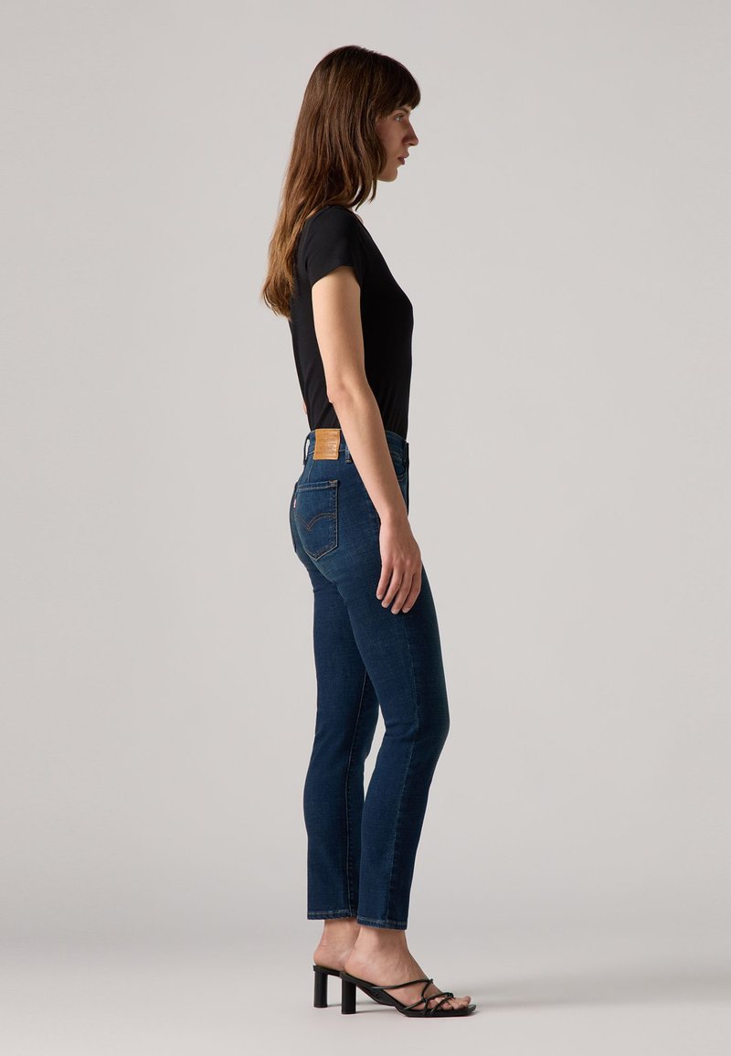 Hoch-taillierte, slim-fit dunkelblaue Jeans aus Denim mit einem Leder-Patch auf der Rückseite, kombiniert mit einem schwarzen eng anliegenden T-Shirt und schwarzen Absatzsandalen.