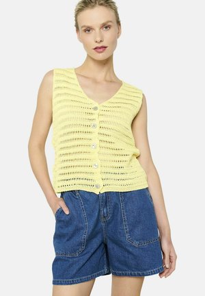 Gelber gestrickter Pullunder mit V-Ausschnitt, ausgestattet mit einer Knopfleiste und offenem Muster, kombiniert mit blauen Denim-Shorts.