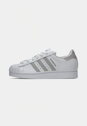 Weißer Adidas Superstar Sneaker mit grauen Streifen, Gummiaußensohle, weißen Schnürsenkeln und Logo auf der Ferse und Zunge, im Profil gezeigt.