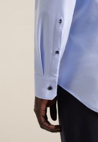 Lichtblauw shirt met een manchet met twee donkere knopen, geborduurde bloemdetails en een gladde textuur.