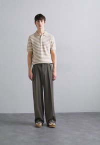 Béžové a krémové pruhované polo tričko, volné tmavé kalhoty a hnědé slip-on boty s bílými akcenty. Model stojí vzpřímeně na neutrálním pozadí.