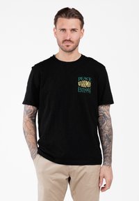 Mężczyzna z tatuażami na ramionach, ubrany w czarną koszulkę "Peace Warriors Est 1994" i beżowe spodnie, stoi z jedną ręką w kieszeni.