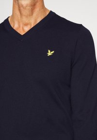 Pull en tricot bleu marine à col en V avec un logo oiseau brodé jaune sur la poitrine, présentant une coupe ajustée et une texture lisse.